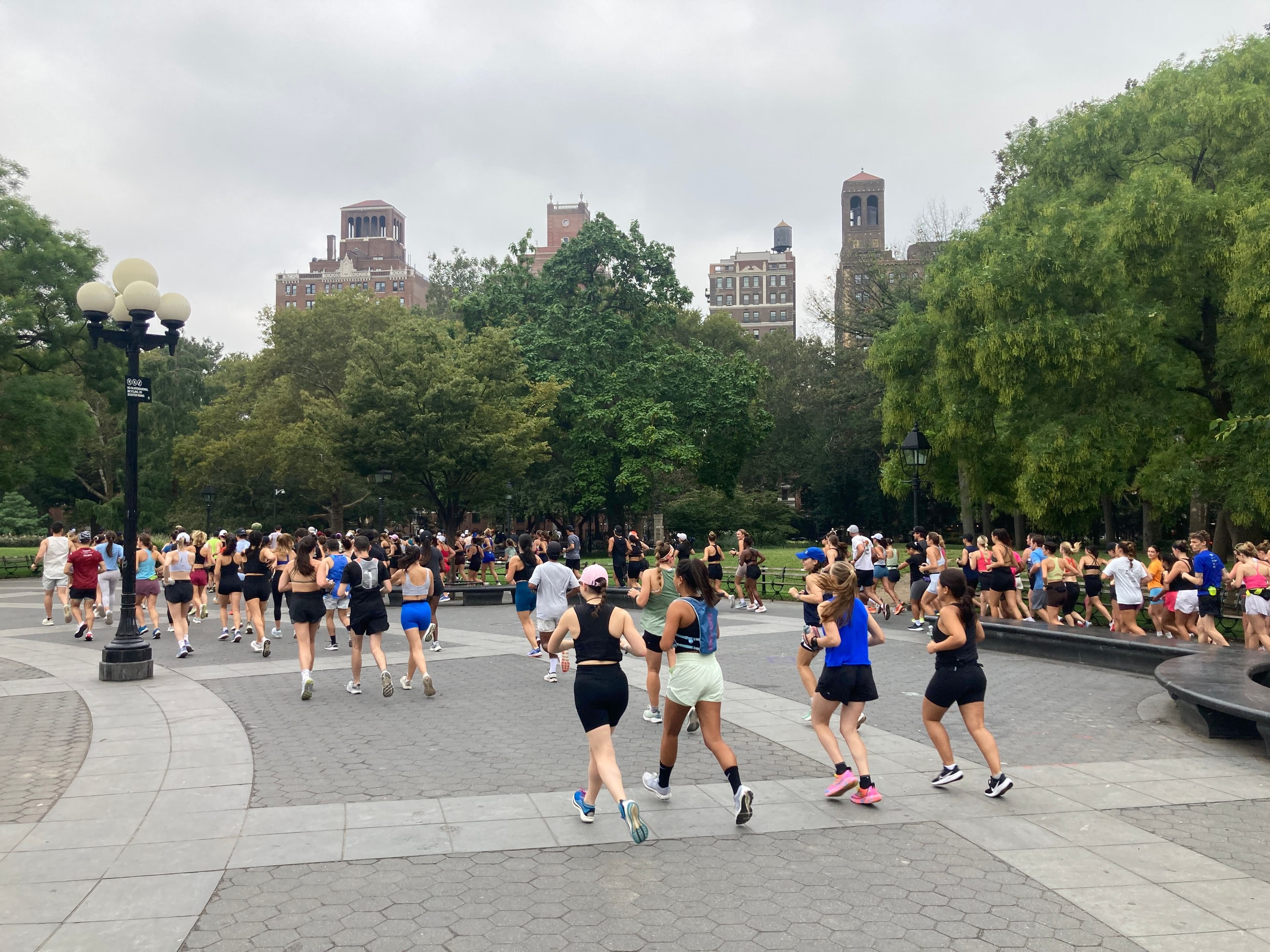 A large group of runners runs together in Washington Square Park.