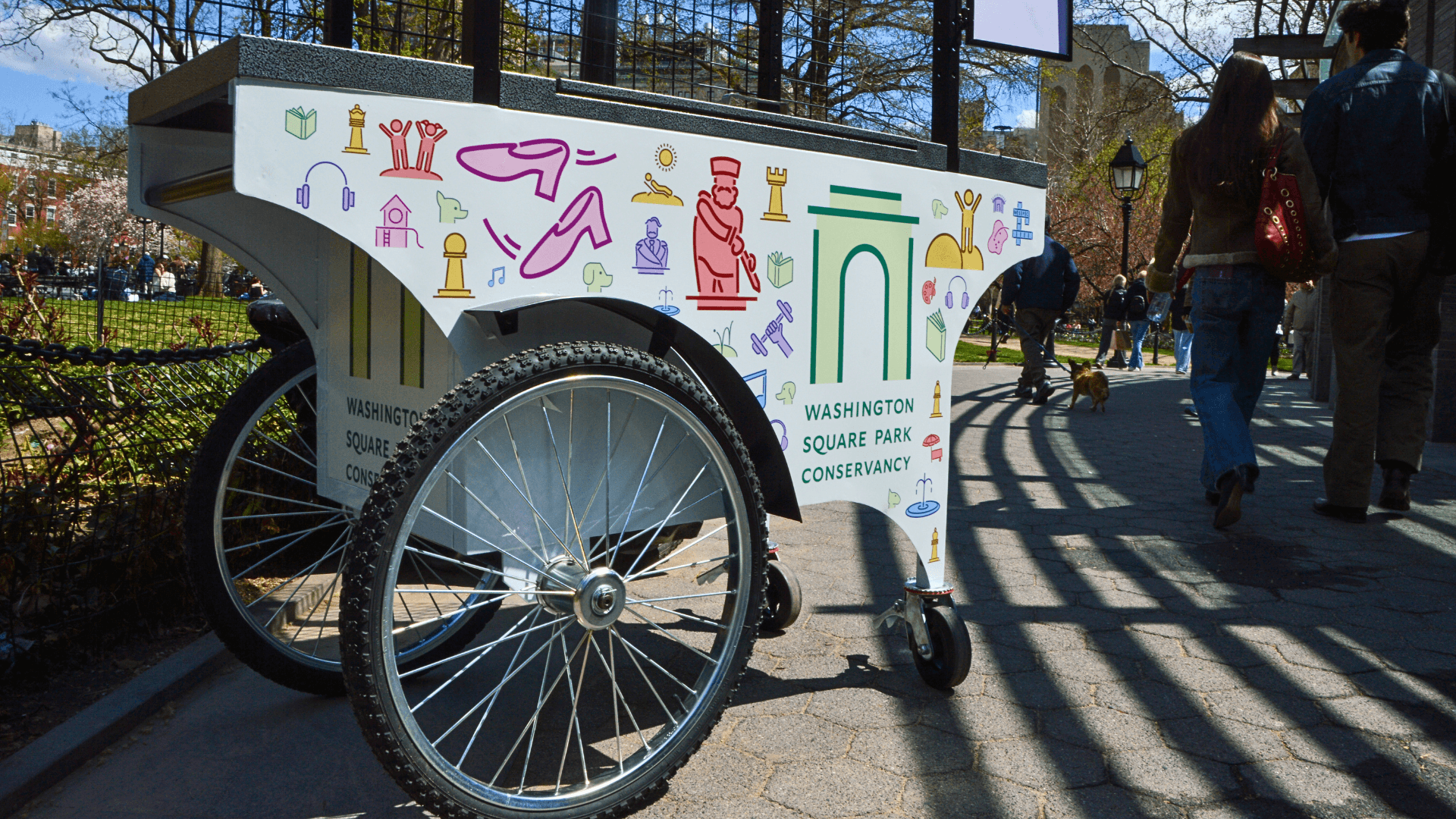 A colorful Washington Square Park Conservancy-brand cart sits in the park while parkgoers pass by.