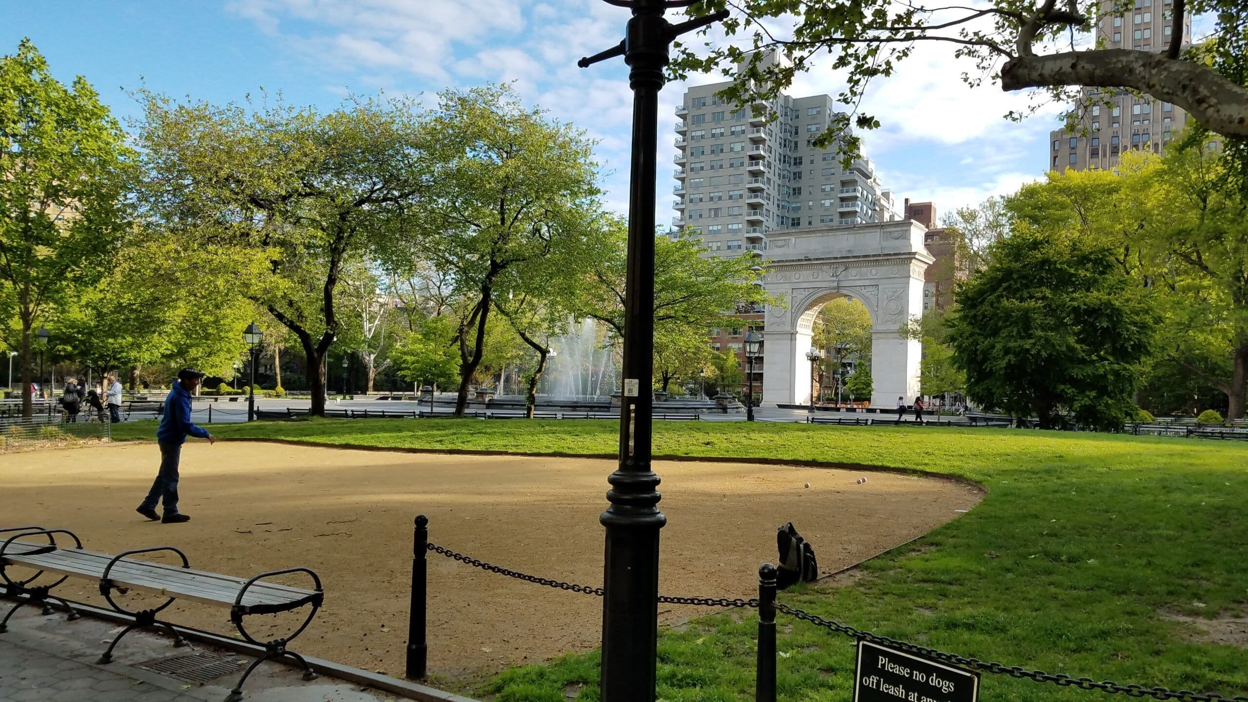 A person uses the pétanque courts in Washington Square Park, with the Washington Square Park Arch behind them.